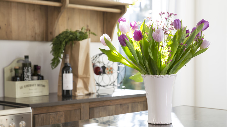 A purple tulip bouquet in a white vase sit on a countertop with wine and groceries in the background.