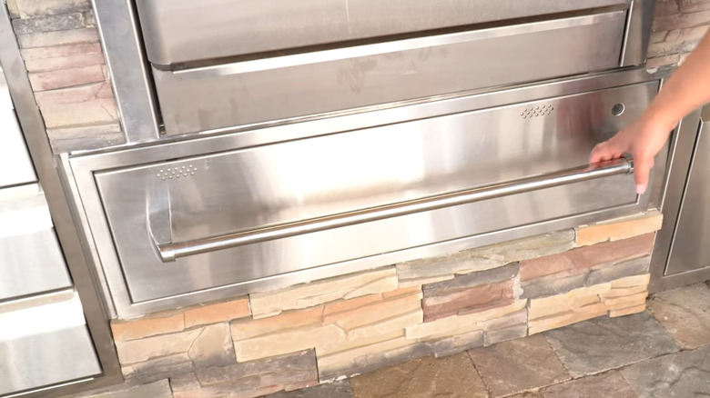 A person opening a stainless steel warming drawer in an outdoor kitchen