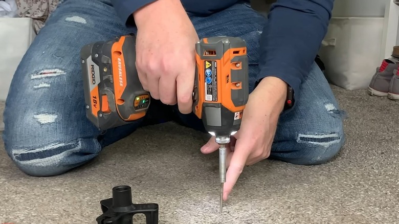 A person uses an electric drill to screw through carpeted floor.