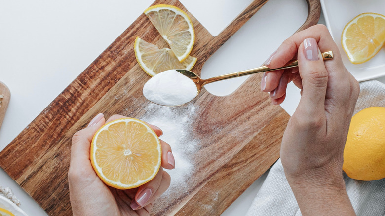 A woman's hands hold a lemon and a spoonful of baking soda to clean wooden board