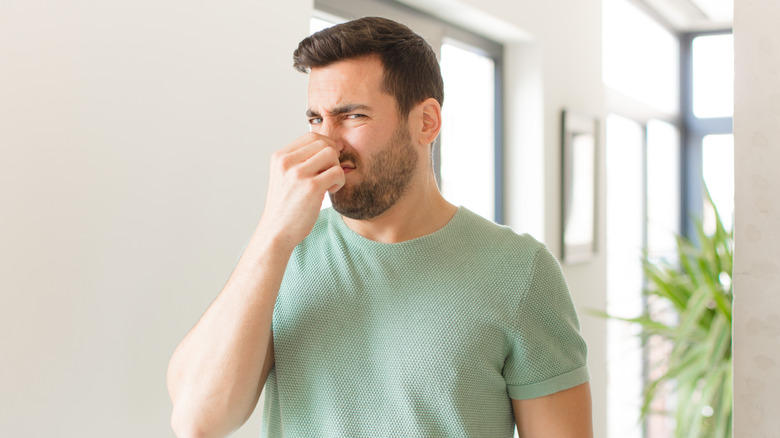 A man holds his nose with a grimace on his face while standing in a house