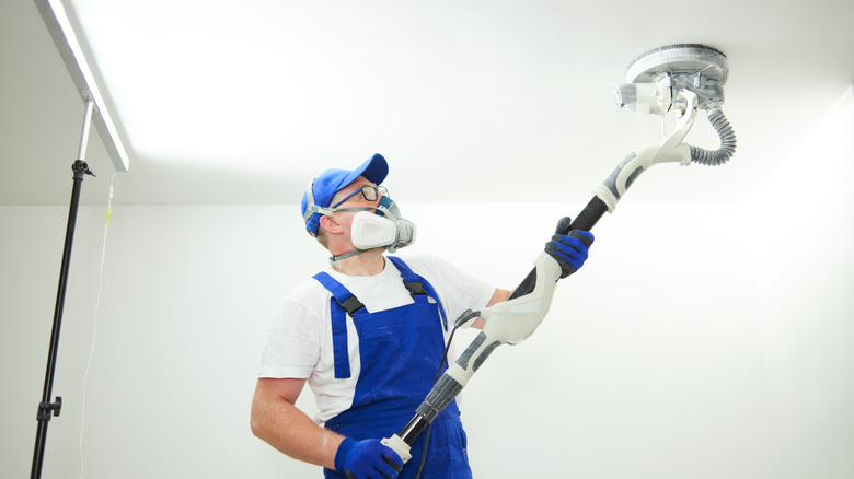 Man using drywall sander on a ceiling with a respirator mask and additional PPE