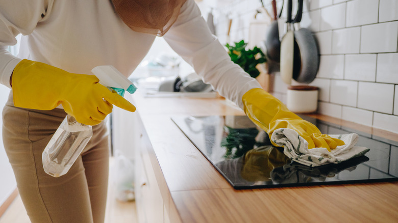 A woman is wearing yellow cleaning goves, holding a spray bottle, and wiping down her glass cooktop with a cloth.