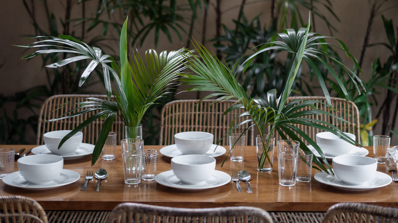 A table set with plates and bowls, and centerpieces of palm fronds in vases.