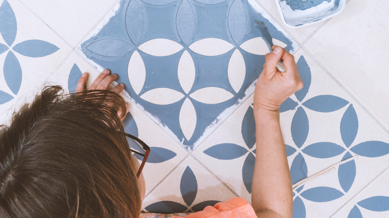 A person stenciling a pattern in blue paint on white tile