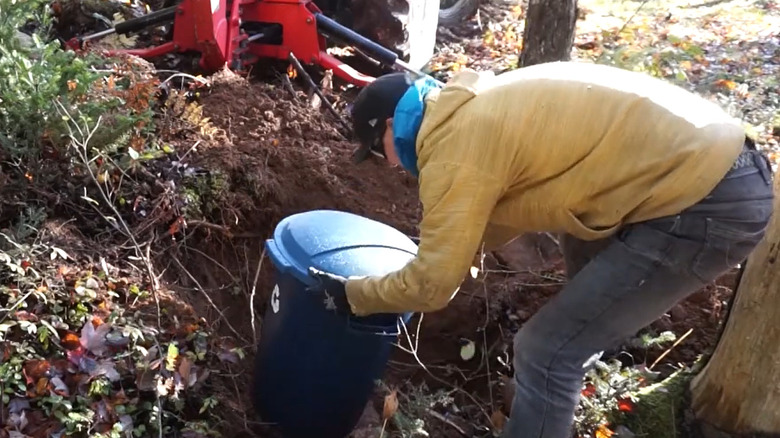 Man placing plastic trash can in hole in ground as a root cellar