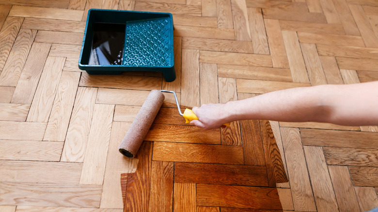 A person applies wood stain to a parquet floor with a roller