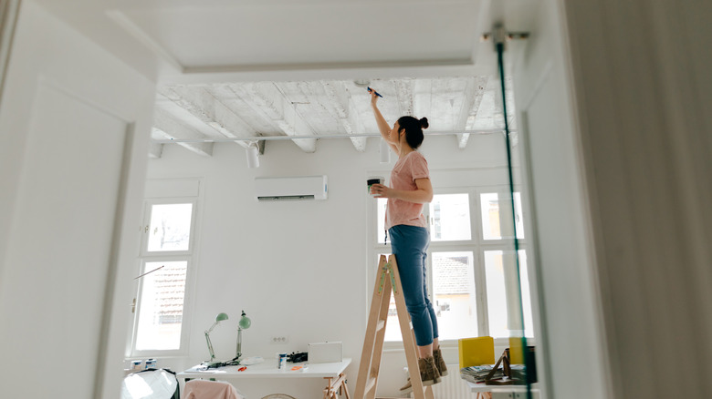 A woman standing on a short ladder is painting her ceiling.