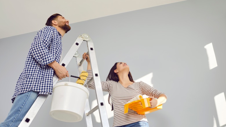 Two people are on a ladder, holding painting equipment and looking toward the ceiling.