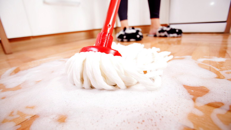 Person mopping wooden floor with a red and white mop, there is some cleaning solution on the floor