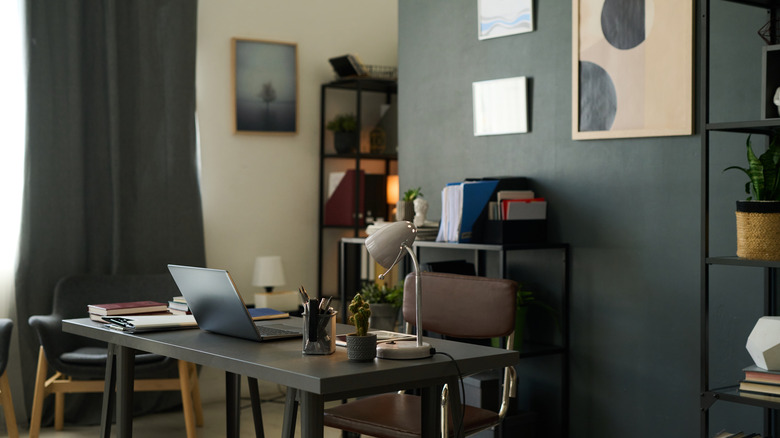 Home office with a dark accent wall, black desk and drapes, wall art, and metal shelving filled with decor