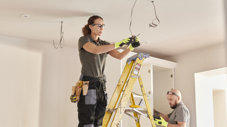 An electrician stands on a ladder to work on wiring in a ceiling.