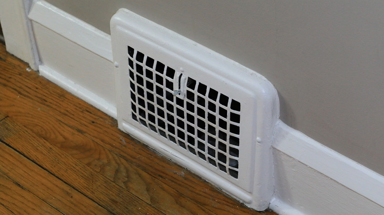 A small air vent, placed low to the gray wall against wooden floor boards.