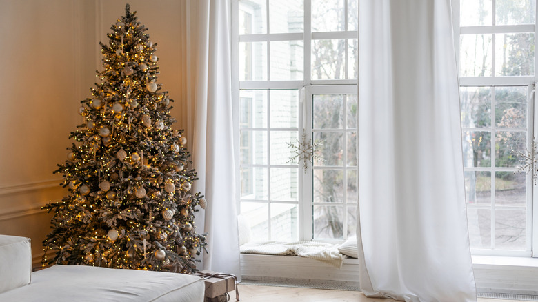 A decorated Christmas tree next to large windows with white curtains