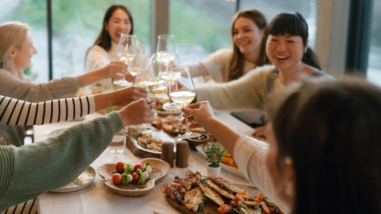 A group of smiling women are sitting around a table clinking their wine glasses together