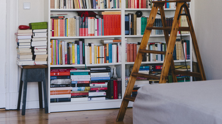 Messy, full white bookshelf in home with stacked books on stool