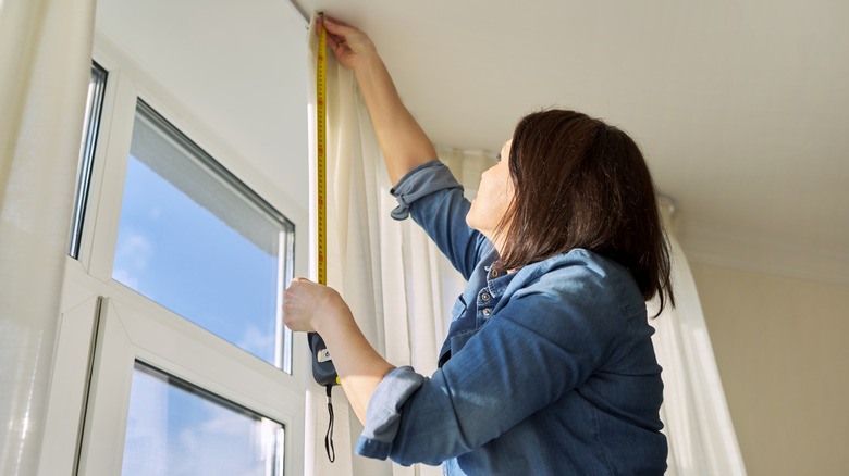 A woman wearing a denim shirt holds steel tape, measuring for curtain length with white sheer curtain beside her