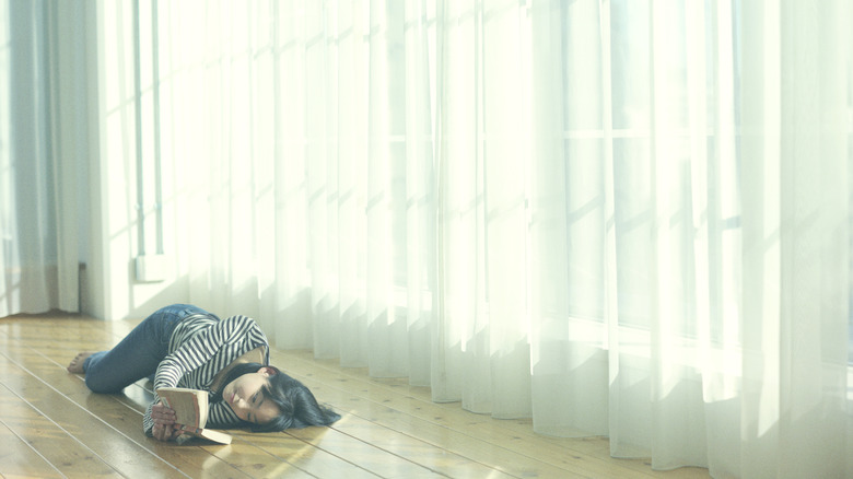 A woman reads a novel on a wood floor beside white sheer curtains kissing the floor with light filtering into room