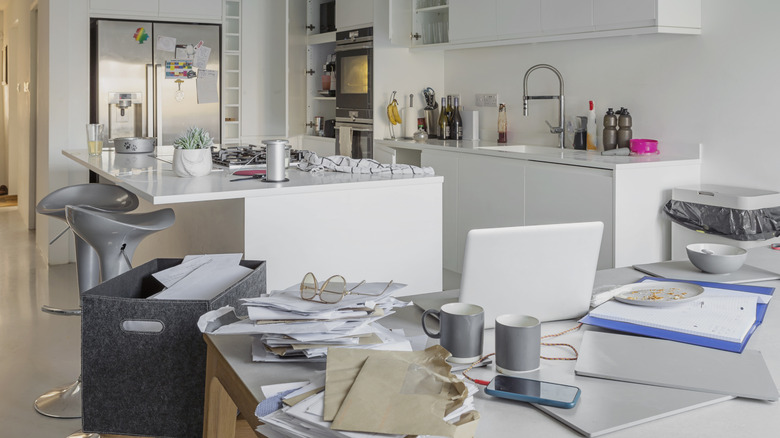 an untidy kitchen and dining area, with papers stacked up, empty mugs, and an open laptop