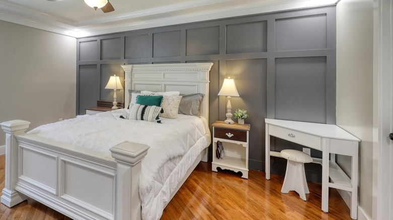 A white bed with two side tables and a desk in the corner rests against a gray board and batten accent wall in a bedroom