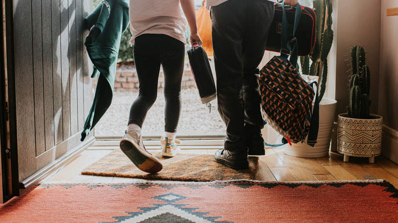 Two kids carrying school bags, exiting entryway with a large rug and doormat.