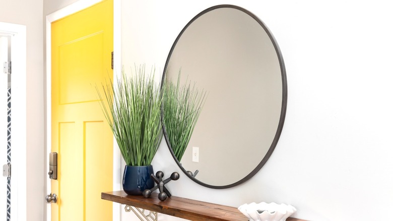 Round mirror over wood shelf holding plant and accessories next to yellow front door