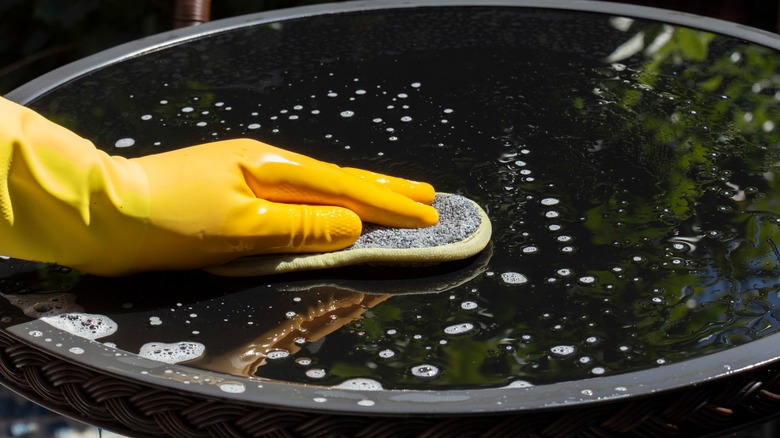 A hand in a marigold glove cleans a glass table