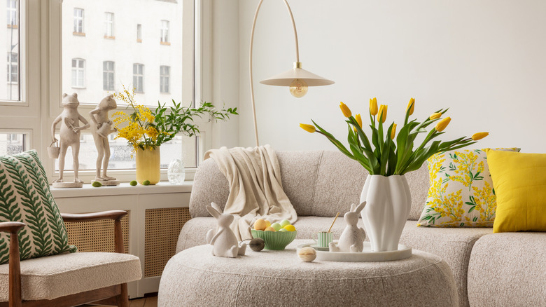 A living room is decorated with a white couch, ottoman, and chair, and white knickknacks with yellow accent pillows and a yellow planter by the window next to frog statues.