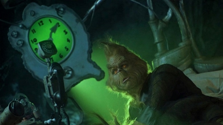 The Grinch sits in bed next to a large alarm clock