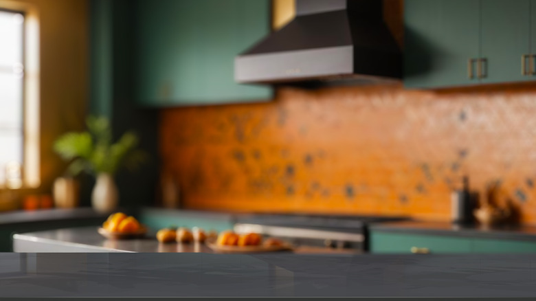 Black marble countertop in foreground with green kitchen cabinets and copper colored backsplash