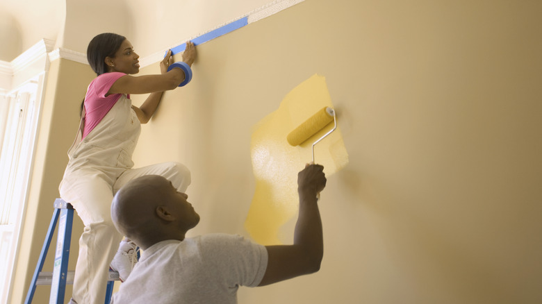 A young couple is painting bright yellow over a beige wall.