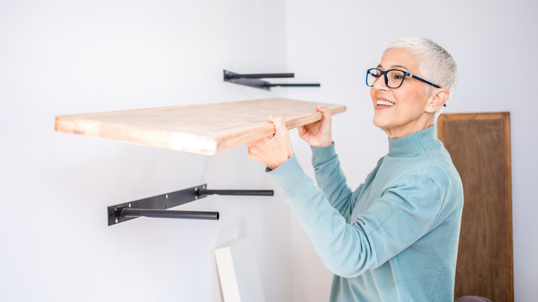 A woman with gray hair installs wooden floating shelves