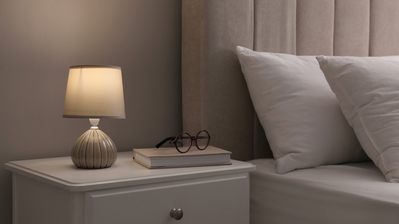 Diffused light coming through small lampshade on white nightstand with a pair of glasses and a book