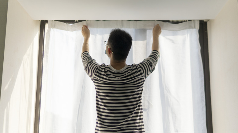 man in front of window hanging white sheer curtains on ceiling track