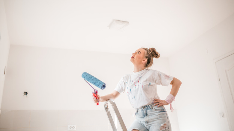 A woman smiles while looking up at a ceiling on a step ladder