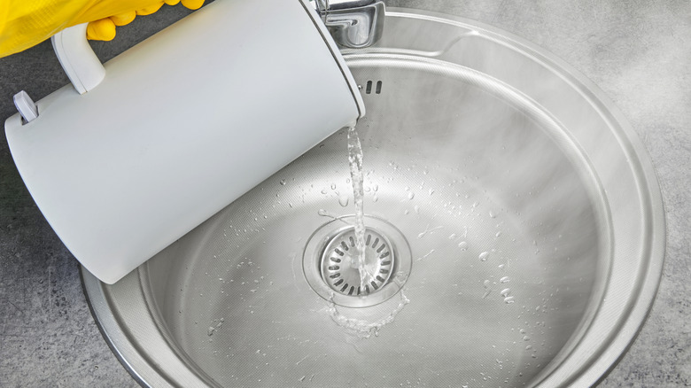 A person in a gloved hand pouring boiling water down a sink drain