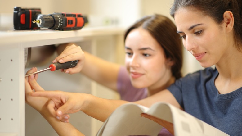 One woman reads an instruction manual while another uses a screwdriver to remove a cabinet door