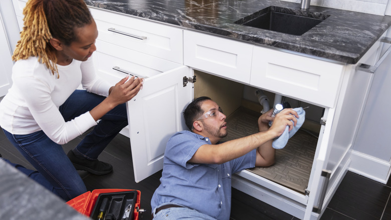 A woman holds a kitchen cabinet open while a man removes a garbage disposal