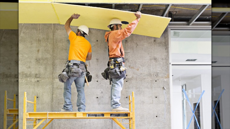 Two workers in hardhats stand on scaffolding while lifting yellow foam board insulation into a ceiling