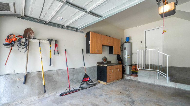 A variety of garden tools hang next to a small cabinet and hot water tank in a garage