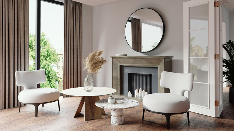 Two white round coffee tables of different heights positioned between two white chairs in front of a gray fireplace with a round mirror over top and a large floor to ceiling window behind one chair with gray drapes