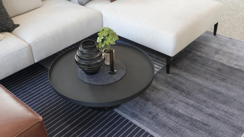 A black round coffee table with a black base and small green plant positioned off center in the corner of a white L-shaped sofa over a gray rug and blue striped rug with a brown leather piece of furniture on the opposite side.