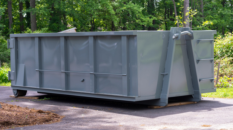 A gray construction dumpster with trees in the background