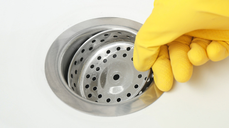 Hand with yellow rubber gloves placing drain strainer in ceramic kitchen sink.