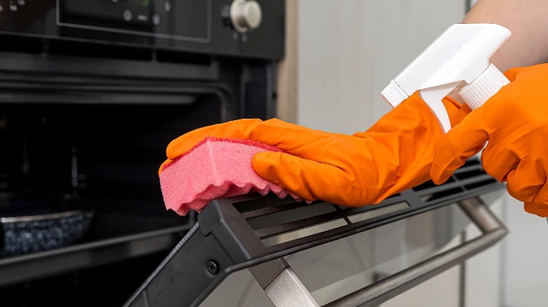Orange gloved hands holding pink sponge on oven door