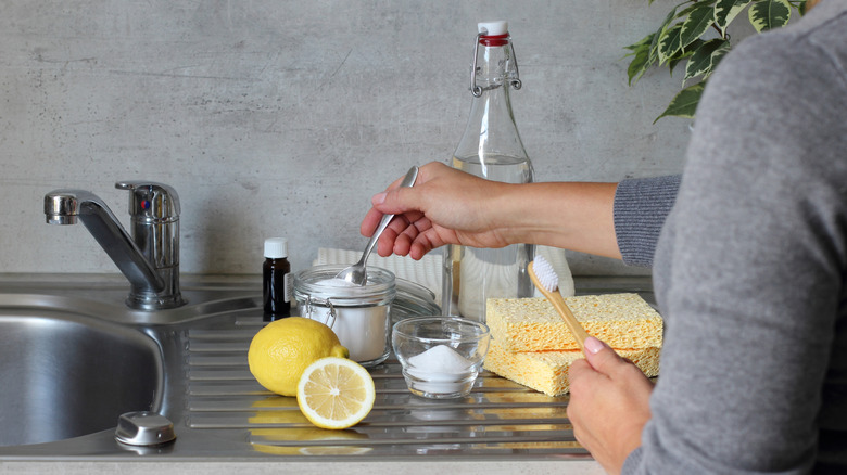 A person holding a spoon and toothbrush near natural cleaning ingredients