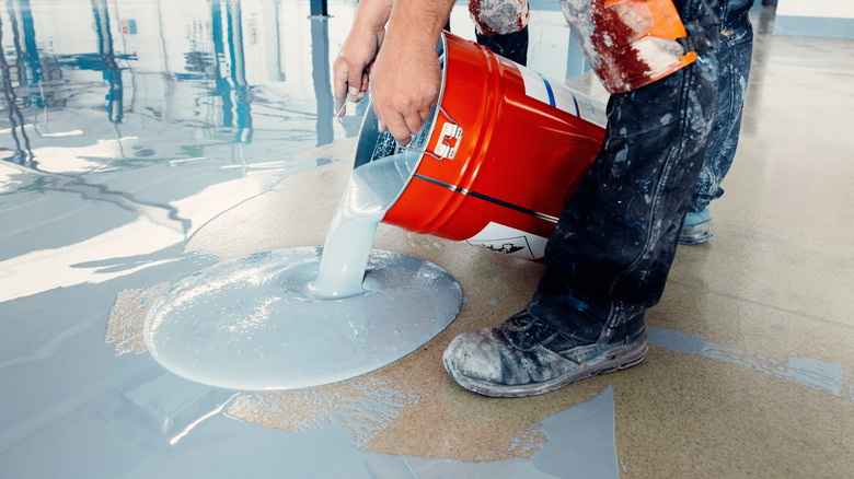 A construction worker pours gray epoxy resin onto a subfloor.