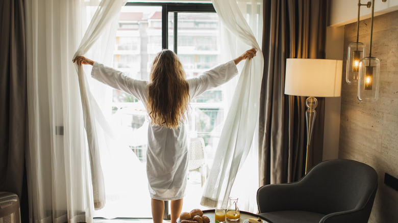 woman standing at window opening curtains