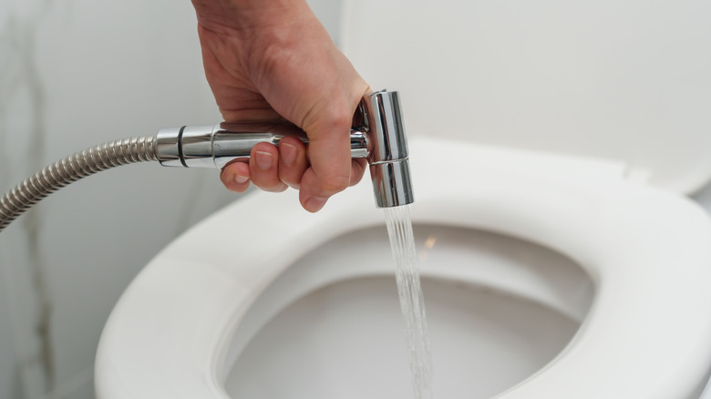 A person is using a handheld bidet to clean the inside of a toilet bowl