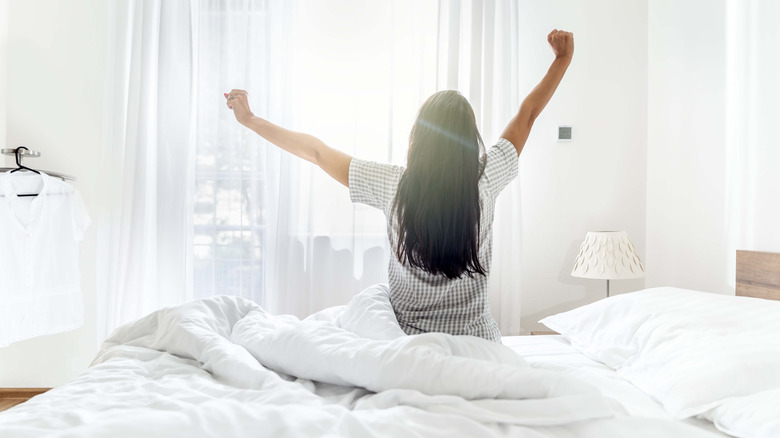 A woman stretches her arms out while sitting on a bed in a sunlight filled room